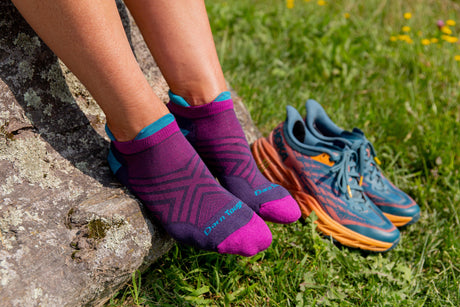 Purple socks with blue accents worn by a person sitting on a rock with running shoes beside them on grass.