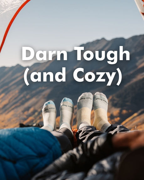 Two pairs of feet wearing socks with text 'Darn Tough (and Cozy)' against a mountainous background.