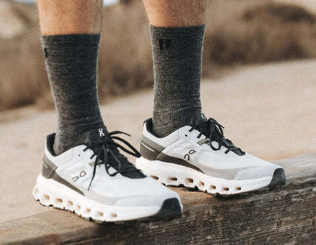 White and gray On running shoes worn with dark gray Hollow crew socks, shown on a person standing on a weathered wooden log outdoors.