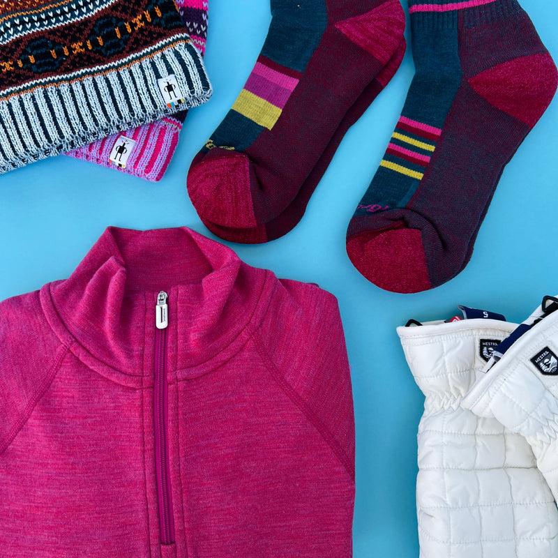 A flat lay of gifts for women, including a pair of socks, mittens, hats, and zip-top base layer 